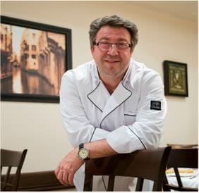 Chef Angelo D in his white chef’s jacket, leaning on a chair in the dining room