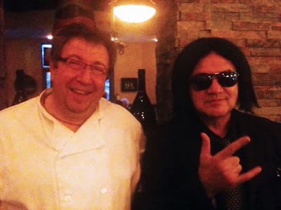 The chef in a white jacket smiling beside a guest in dark glasses