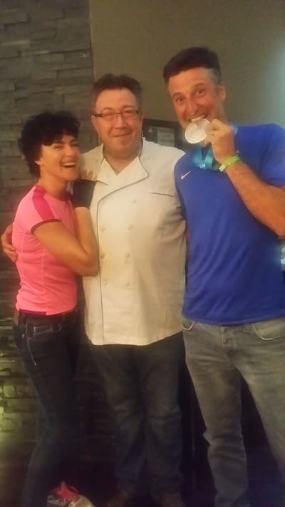 The chef laughing with the same two guests; one bites a medal