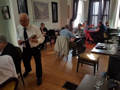 A man in a white shirt and tie carries menus through the dining room while guests sit at tables