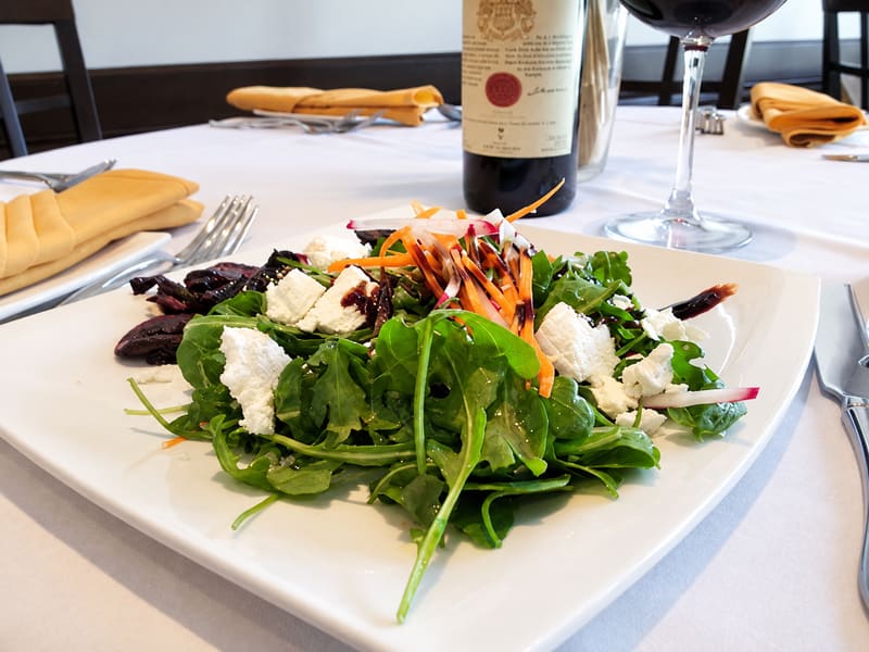 A salad of greens and white cheese on a square plate, with a bottle of red wine behind